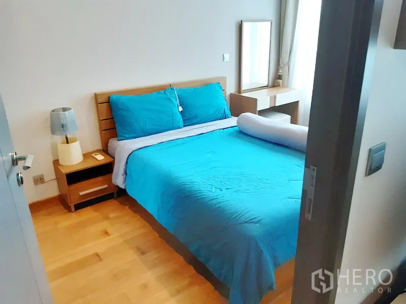 Condominium for rent in Khlong Toei, Bangkok - Cozy bedroom with double bed, wood flooring, bedside table and vanity by a large window.