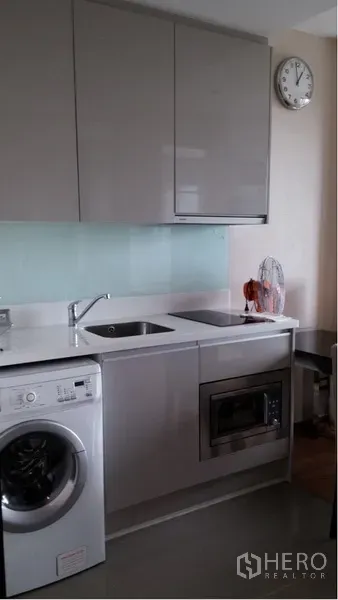 Condominium for rent in Ratchathewi, Bangkok - Compact modern kitchen with white countertop, sink, wall cabinets, microwave, and front-load washing machine.