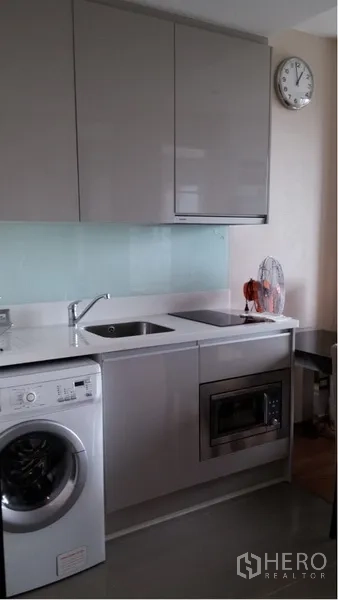 Condominium for rent in Ratchathewi, Bangkok - Compact modern kitchen with white countertop, sink, wall cabinets, microwave, and front-load washing machine.