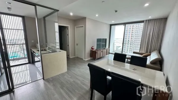 Condominium for rent - Open living zone showing glass-enclosed kitchen, dining table, sofa, TV, and floor-to-ceiling windows.