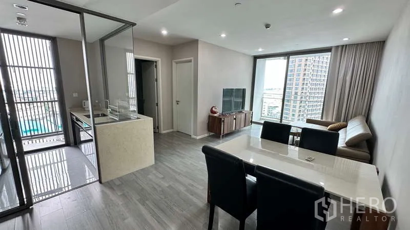 Condominium for rent - Open living zone showing glass-enclosed kitchen, dining table, sofa, TV, and floor-to-ceiling windows.