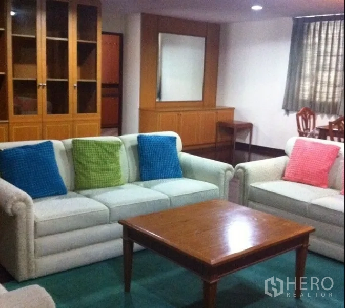 Condominium for rent in Watthana, Bangkok - Living area with cushioned sofas, glass-front cabinets, and a dining space beyond.
