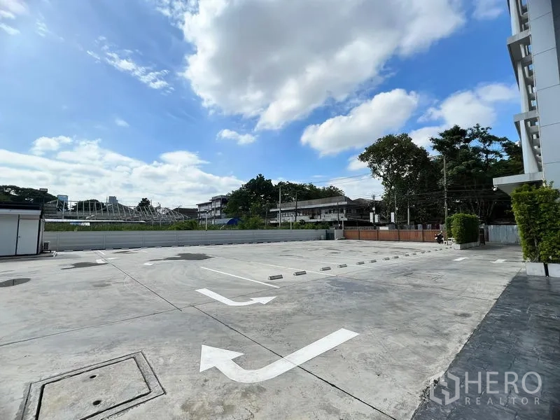 零售空间 出租在Huai Khwang, Bangkok - 宽敞的室外停车场，车位与指引箭头清晰，邻近现代低层建筑与蓝天白云。