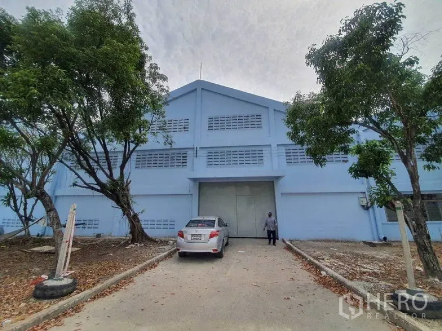 货栈 出租在Nakhon Pathom - 蓝色仓库正面，宽大装卸门、车道与遮荫树。