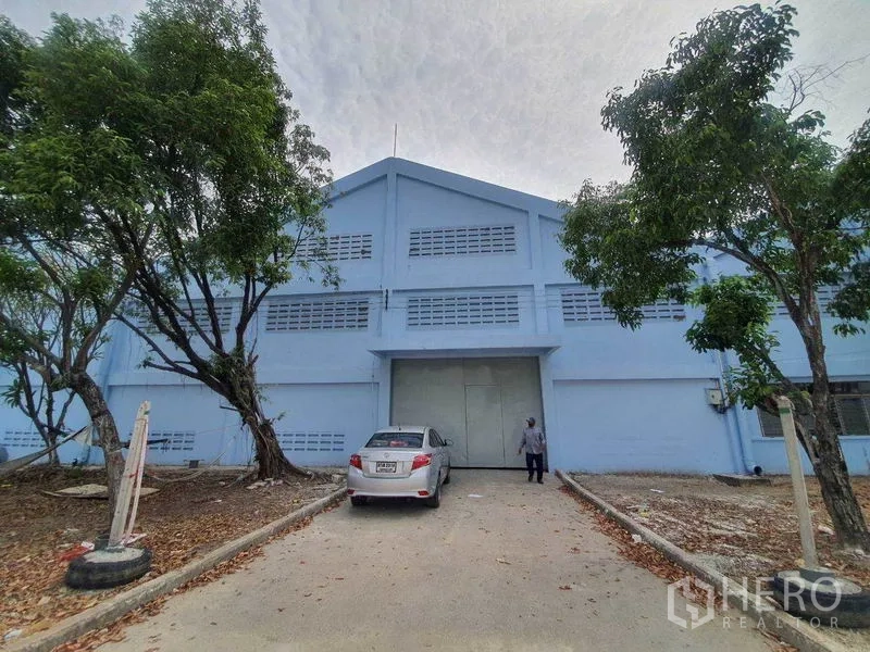货栈 出租在Nakhon Pathom - 蓝色仓库正面，宽大装卸门、车道与遮荫树。