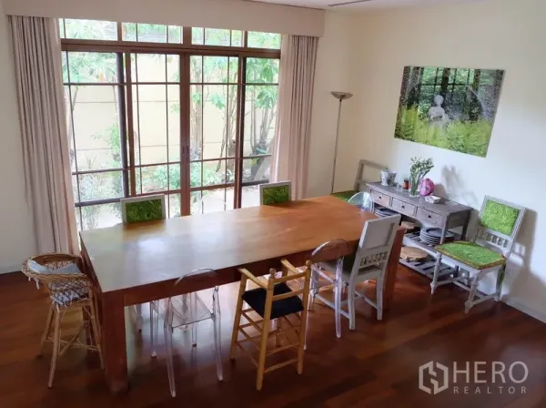 Detached House for rent in Watthana, Bangkok - Dining area with a long wooden table, mix-and-match chairs, and floor-to-ceiling windows facing greenery.