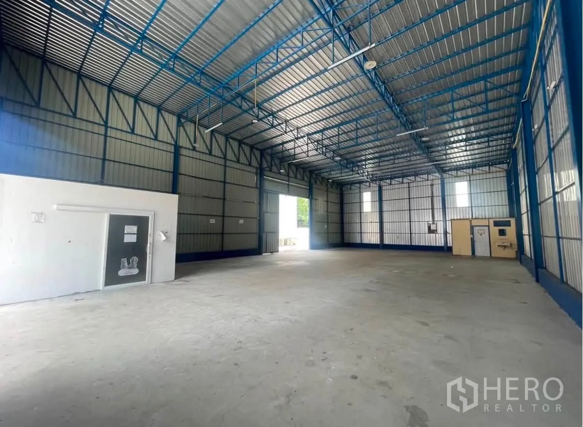 Warehouse for rent in Wang Thonglang, Bangkok - Spacious warehouse interior with a high steel truss roof, concrete floor, and a small enclosed room.