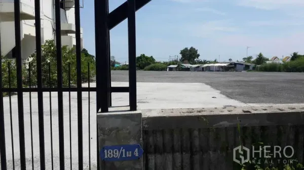 Shophouse for rent in Bang Bo, Samut Prakan - Gated entrance opening to a wide hardstand yard beside greenery.