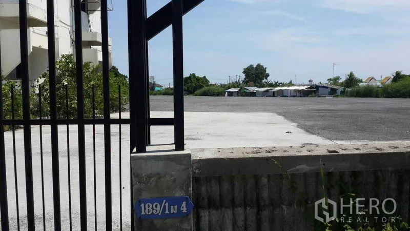 Shophouse for rent in Bang Bo, Samut Prakan - Gated entrance opening to a wide hardstand yard beside greenery.