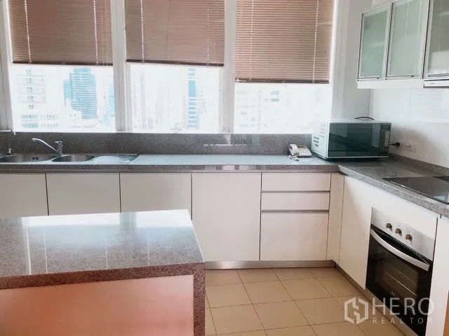 Condominium for rent or sale in Khlong Toei, Bangkok - Modern white kitchen corner with L-shaped counters, stone worktops, cooktop and city views through wide windows.
