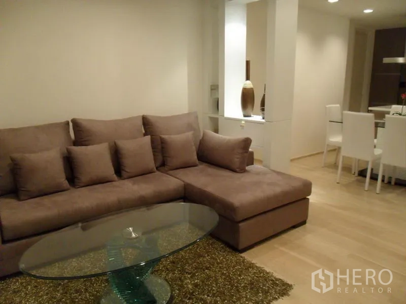 คอนโด ให้เช่าในKhlong Toei, Bangkok - โซฟา L‑shape พร้อมหมอน โต๊ะกลางกระจกวงรีบนพรม ในห้องนั่งเล่นโทนอบอุ่น