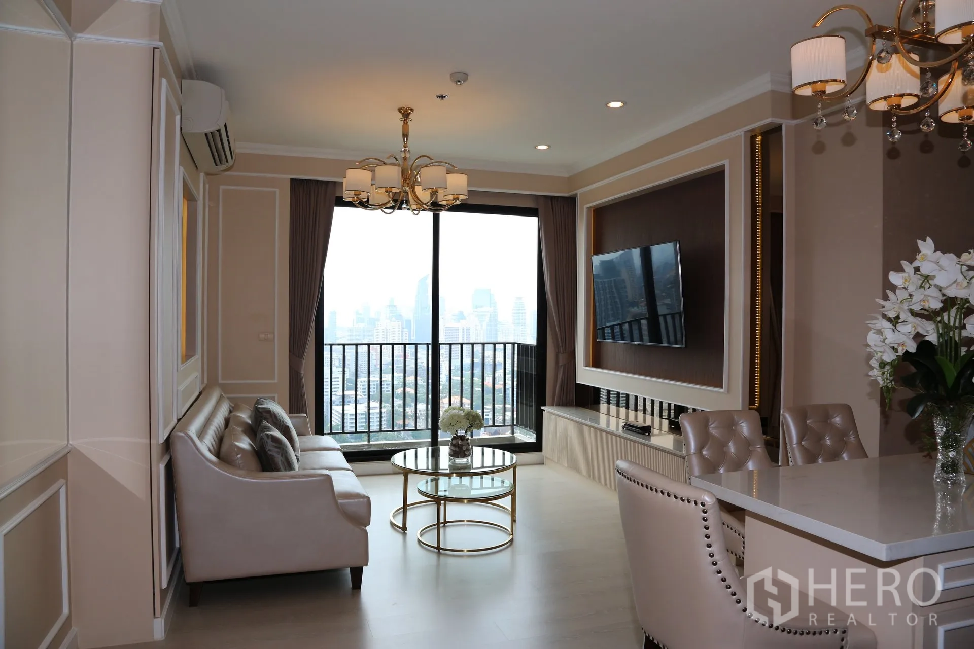 Condominium for rent in Huai Khwang, Bangkok - Open-plan lounge and dining area with city-view balcony, wall-mounted TV, marble-top dining island, upholstered chairs and chandeliers.