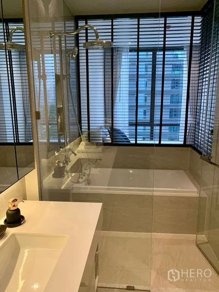 Condominium for rent in Pathum Wan, Bangkok - Modern bathroom with glass shower, bathtub, and large window with blinds.