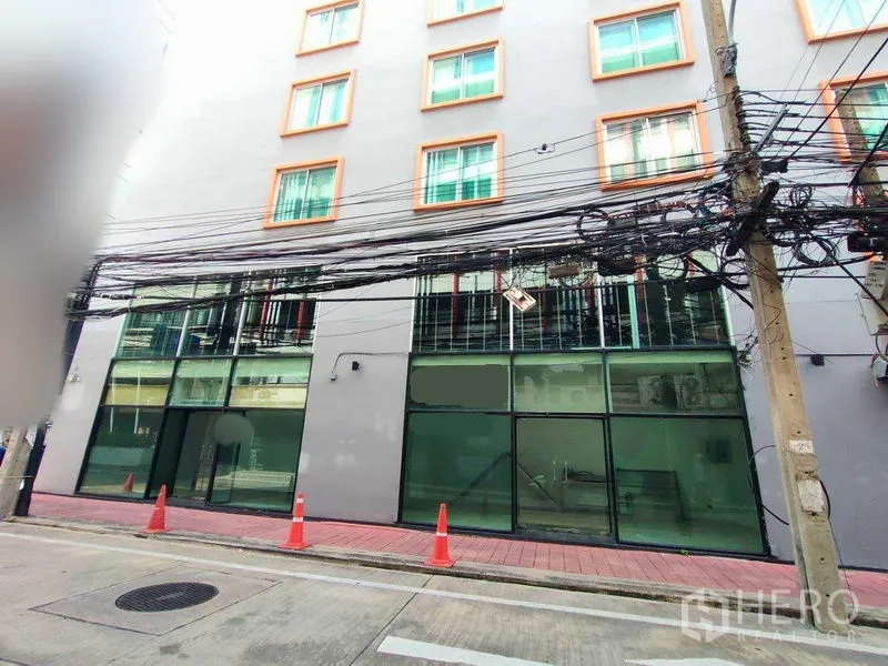 พื้นที่ขายปลีก ให้เช่าในWatthana, Bangkok - หน้าร้านค้าชั้นล่างกระจกเต็มบานใต้ตึกสูงกลาง ทางเท้าปูอิฐสีแดง มีกรวยจราจร และสายไฟเหนือศีรษะ