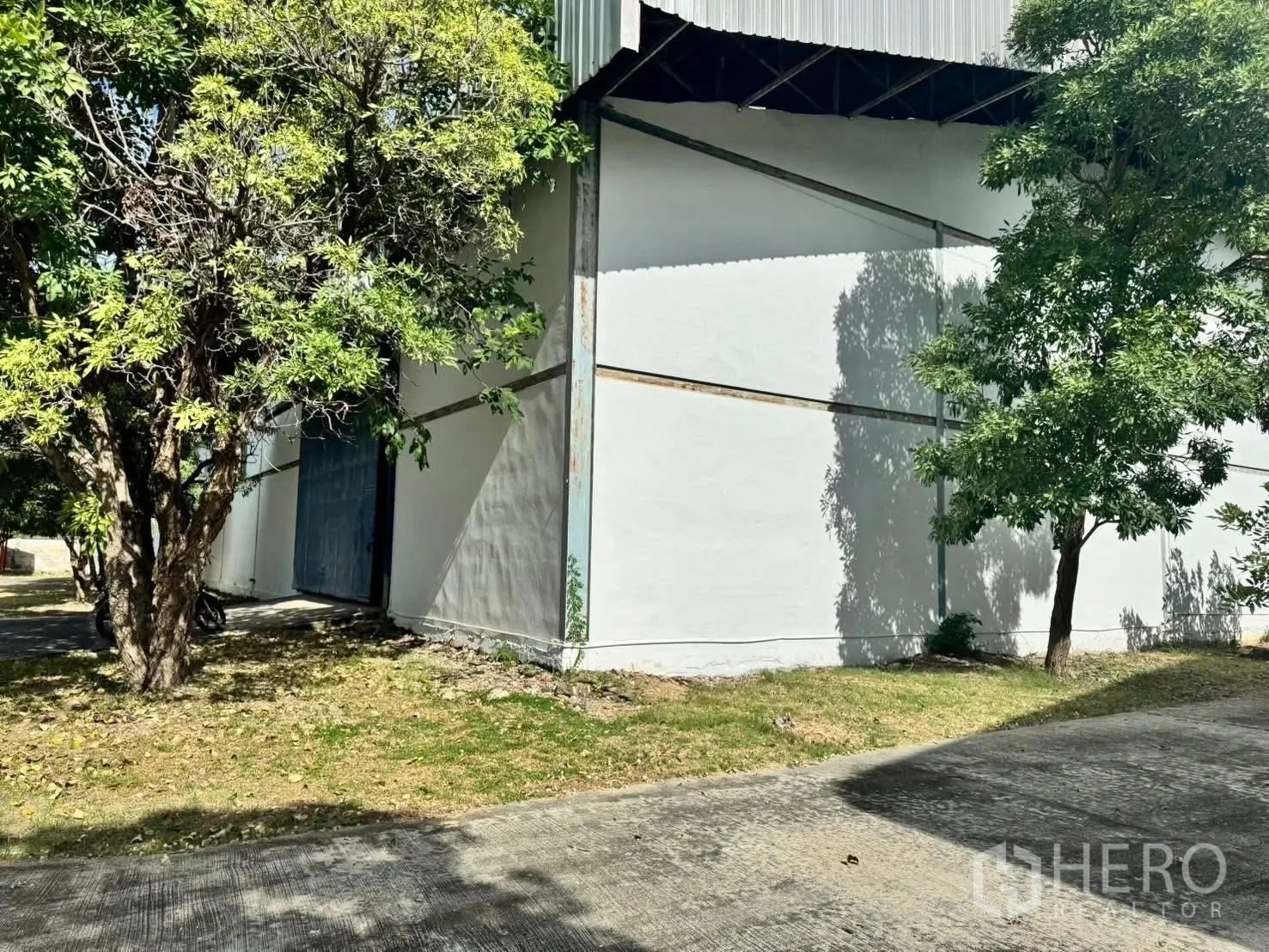 Factory for rent in Bang Phli, Samut Prakan - Side view of the factory showing white walls, blue door, trees, and paved path.