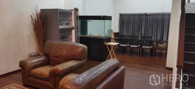 Townhouse for rent in Khlong Toei, Bangkok - Open‑plan living and dining area with leather sofas, a large aquarium, shelving, and a staircase.