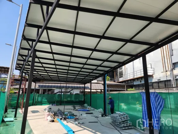 零售空间 出租在Khlong Toei, Bangkok - 带黑色钢柱与大跨度屋顶的覆盖式商铺区，混凝土地面摆放施工材料，城市环境。