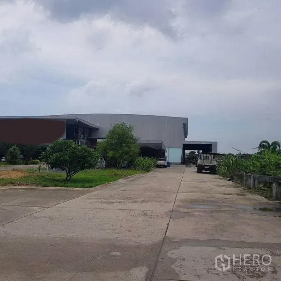 Home Office for sale in Khlong Luang, Pathum Thani - Entrance approach to a large warehouse compound with greenery on both sides.