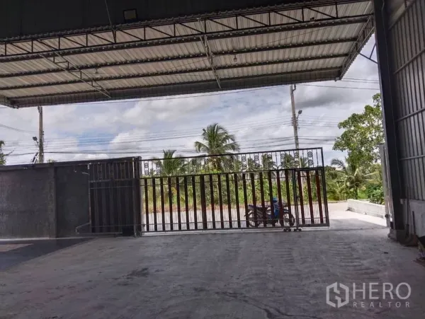 Warehouse for sale in Sai Noi, Nonthaburi - Covered loading bay with wide metal gate opening to the road and greenery.