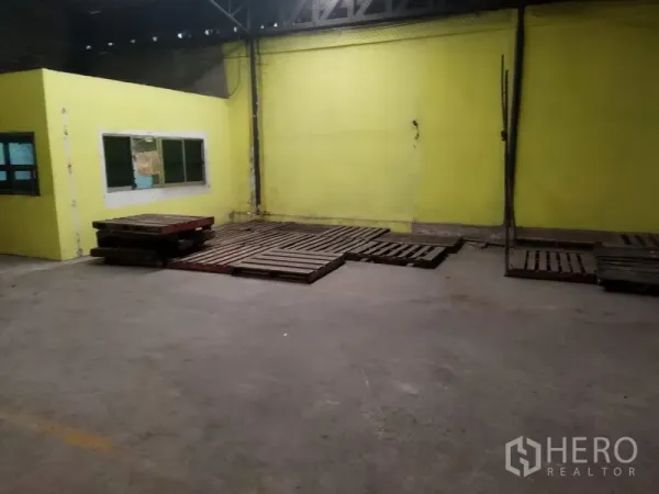 Warehouse for rent in Khlong Sam Wa, Bangkok - Warehouse corner featuring a yellow internal office with windows and stacked wooden pallets on a concrete floor.