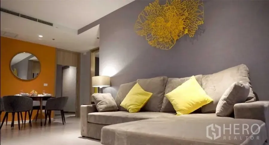Condominium for rent in Pathum Wan, Bangkok - Living room with large plush sofa, yellow cushions and decorative wall sculpture.