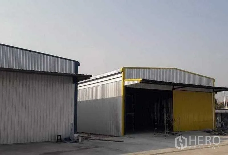 货栈 出租在Khlong Sam Wa, Bangkok - 侧视双体金属仓库，黄色立面与遮雨棚，旁有车道及装卸区。