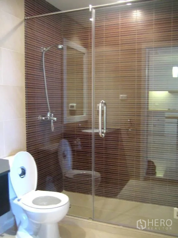 Condominium for rent in Ratchathewi, Bangkok - Bathroom with glass-enclosed shower, brown mosaic wall tiles and white toilet.