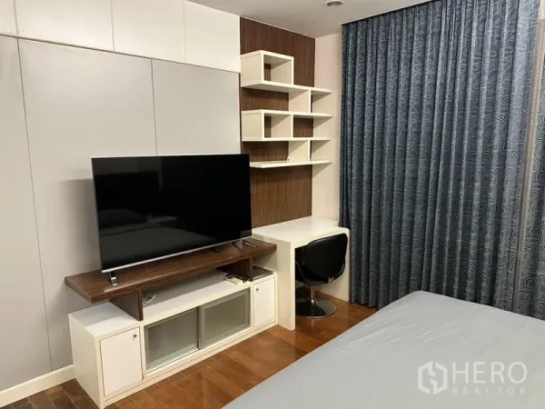 Condominium for rent in Watthana, Bangkok - Bedroom corner with TV console, built-in desk and geometric shelves, wood floors and blackout curtains.