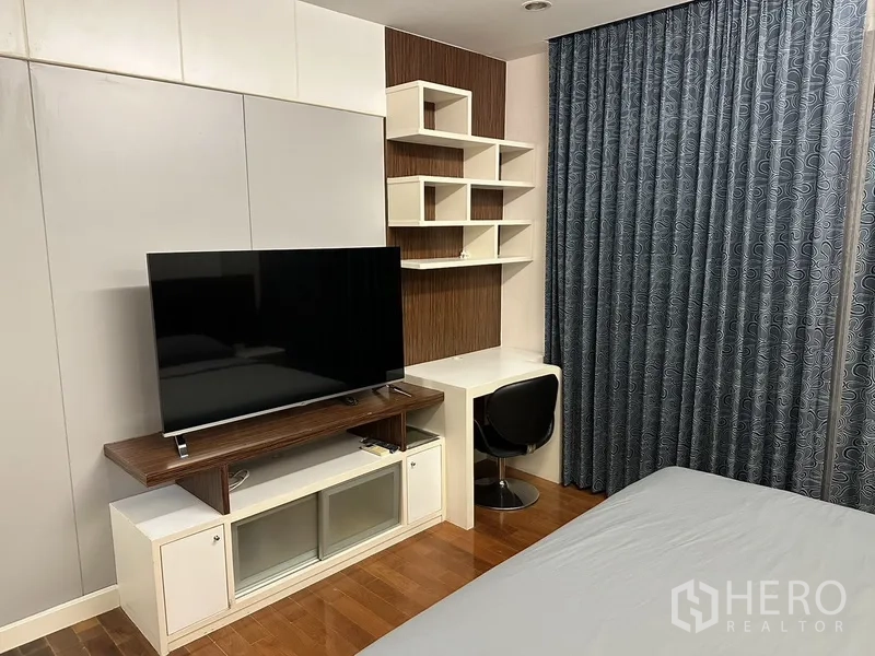 Condominium for rent in Watthana, Bangkok - Bedroom corner with TV console, built-in desk and geometric shelves, wood floors and blackout curtains.