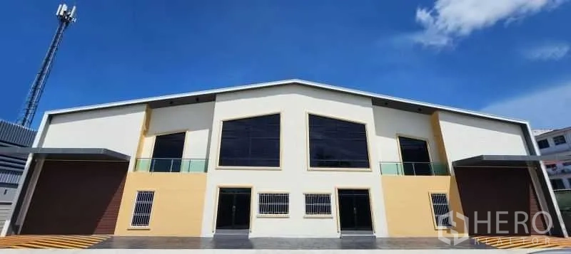 Warehouse for rent in Bang Bua Thong, Nonthaburi - Modern twin-bay warehouse exterior with two roller shutters, glass panels, and a parking apron under a blue sky.