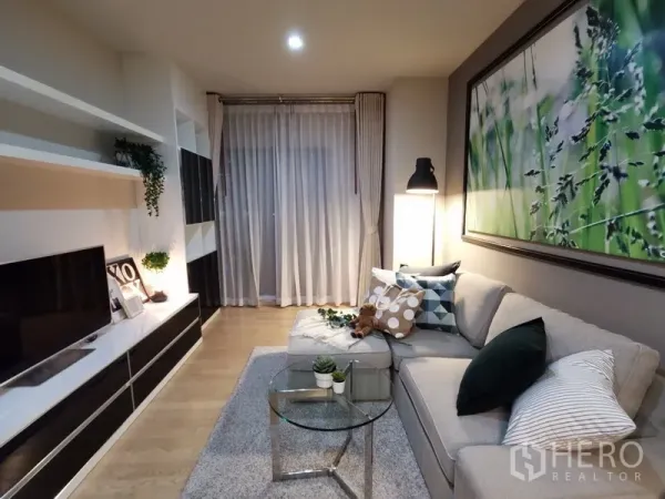 公寓 出售在Bangkok - 舒适客厅，L型沙发、内置电视墙、纱帘与柔和灯光。