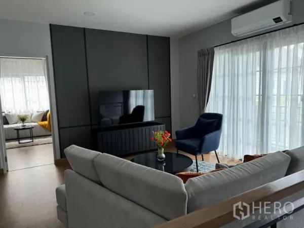 Detached House for rent or sale in Bang Phli, Samut Prakan - Upstairs family lounge with sectional sofa, TV wall unit, blue armchair and sheer curtains.