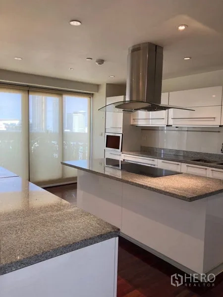 Condominium for rent in Bang Rak, Bangkok - Modern white kitchen with island, granite counters, and stainless range hood opening to a balcony.