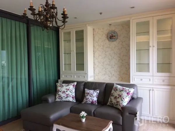 คอนโด ให้เช่าในKhlong Toei, Bangkok - ห้องนั่งเล่นอบอุ่น โซฟาหนังสีเทา หมอนลายดอก โคมระย้า วางคู่ตู้กระจกบิลท์อินและผ้าม่านสีเขียว