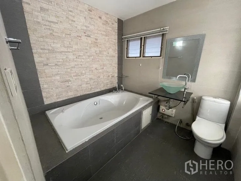 Detached House for rent in Khlong Toei, Bangkok - Bathroom with built-in bathtub, stone accent wall and modern glass-bowl vanity.