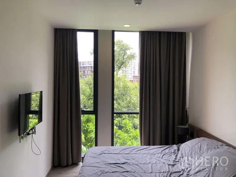 Condominium for rent in Watthana, Bangkok - Bedroom with large windows, blackout curtains, wall-mounted TV, and a treetop view.