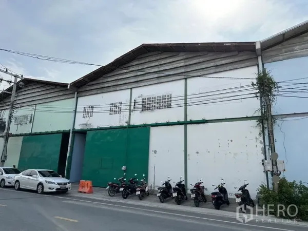 Factory for rent in Bang Bo, Samut Prakan - Street-facing factory facade with large green sliding doors and parked vehicles.