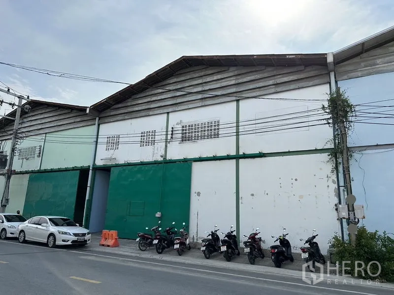 Factory for rent in Bang Bo, Samut Prakan - Street-facing factory facade with large green sliding doors and parked vehicles.
