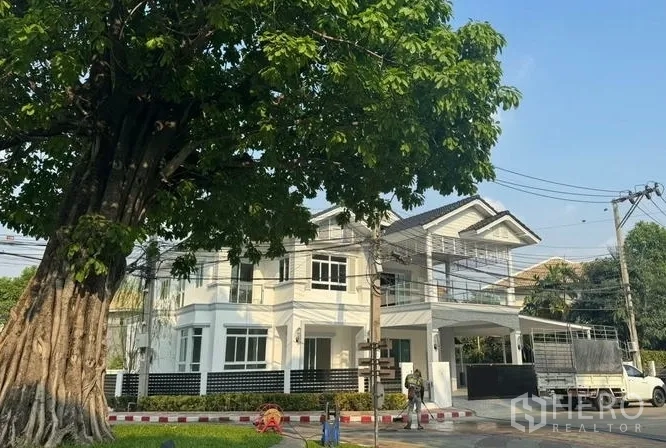 Detached House for sale in Pak Kret, Nonthaburi - Front façade shaded by a large tree, showing balcony, gate and covered parking.