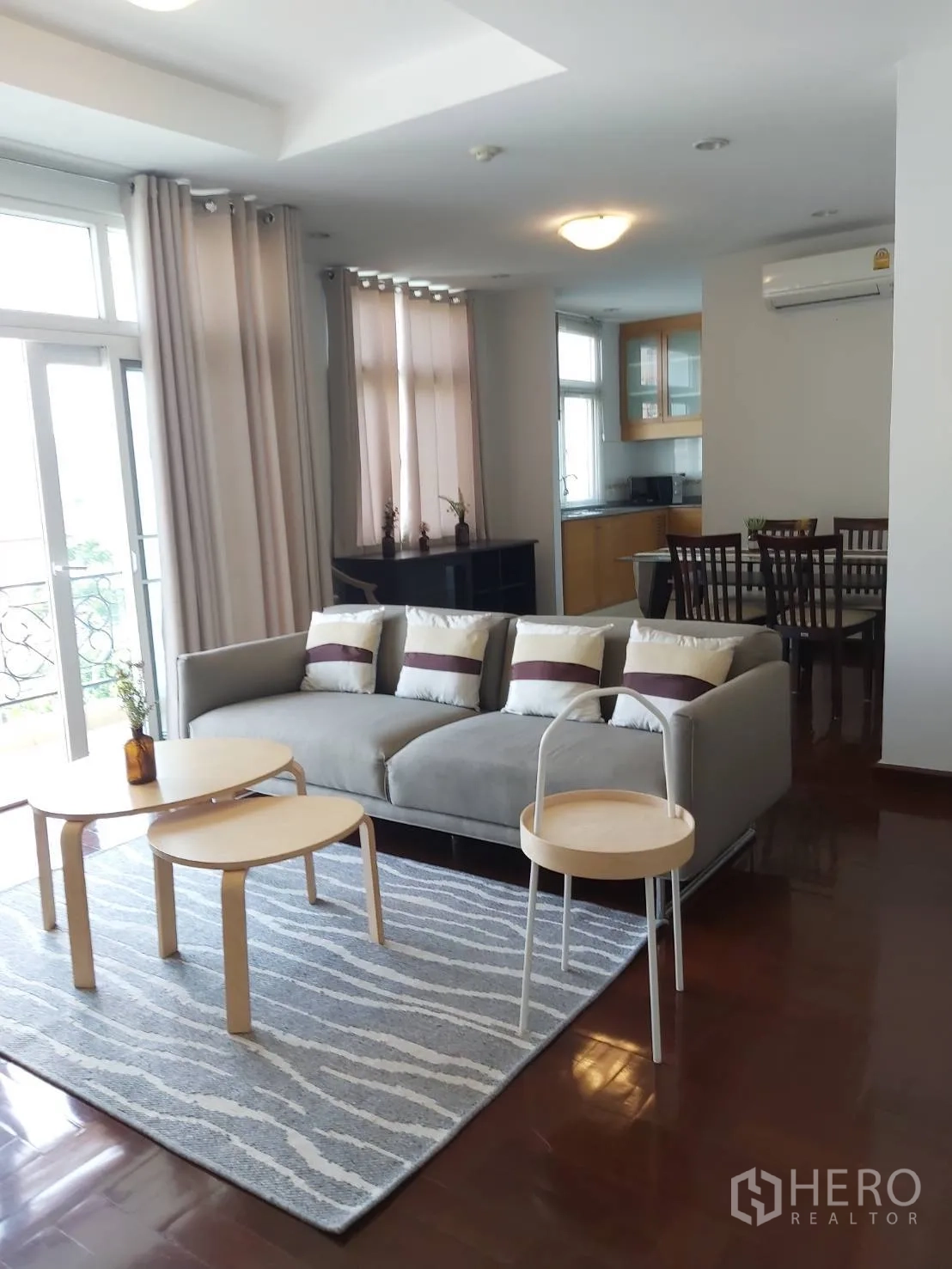 Apartment for rent in Khlong Toei, Bangkok - Bright living room with gray sofa, nesting coffee tables, striped rug, and balcony doors with curtains.