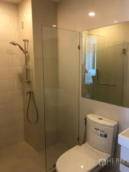 Condominium for rent in Huai Khwang, Bangkok - Modern bathroom with glass shower enclosure, handheld shower, toilet and large mirror.