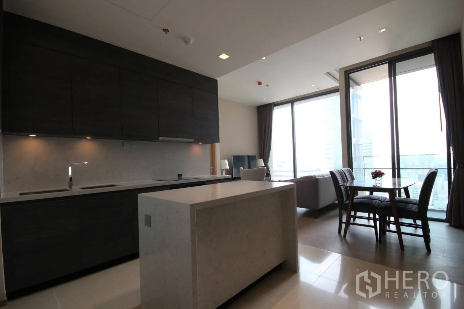 Condominium for rent in Watthana, Bangkok - Contemporary kitchen with dark cabinets, white island, and dining set beside floor-to-ceiling balcony windows.