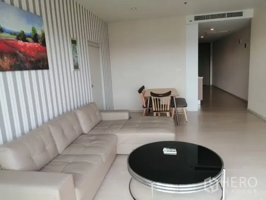 公寓 出租在Watthana, Bangkok - 明亮客厅，米色L形沙发、圆形茶几与条纹特色墙，并设有小型用餐区。