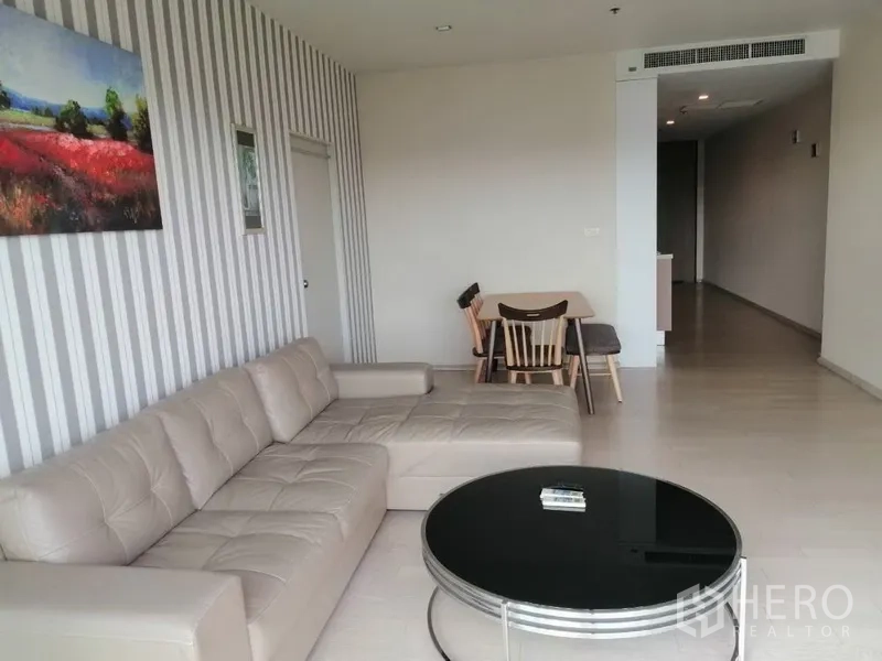 公寓 出租在Watthana, Bangkok - 明亮客厅，米色L形沙发、圆形茶几与条纹特色墙，并设有小型用餐区。