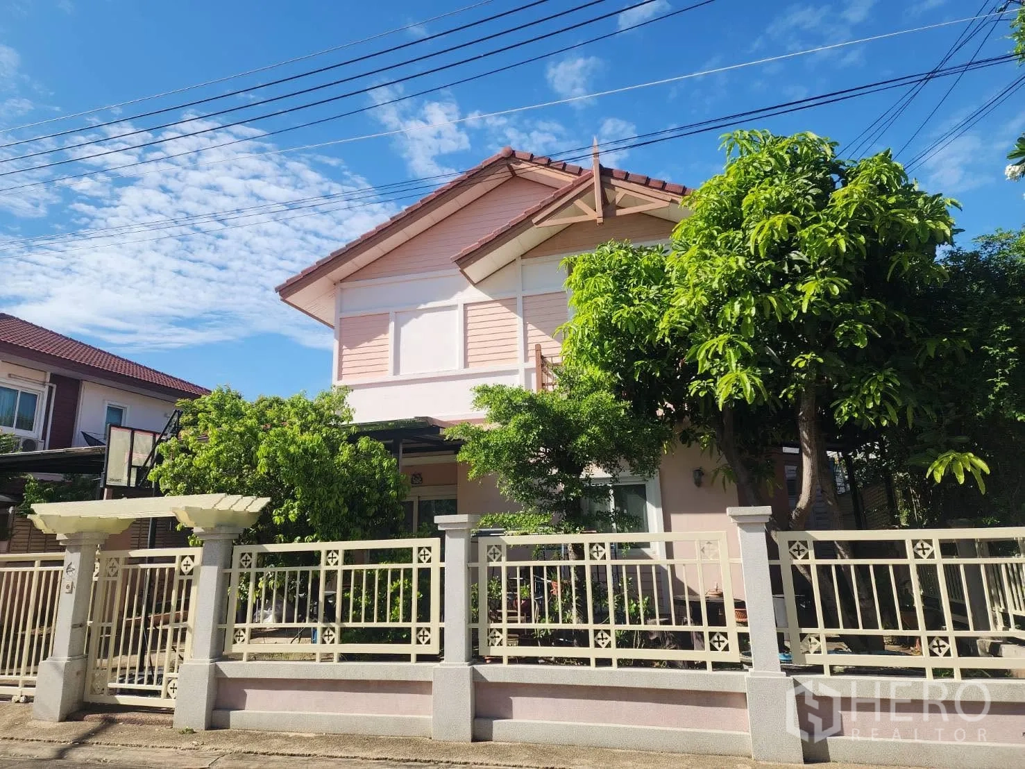 Detached House for rent in Lam Luk Ka, Pathum Thani - Two-storey house exterior with front fence, balcony detail, and mature trees under a blue sky.