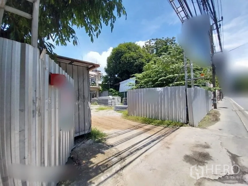 Land for rent in Khlong Toei, Bangkok - Gated land plot with corrugated metal fence, dirt driveway, and roadside access in Khlong Toei under a blue sky.