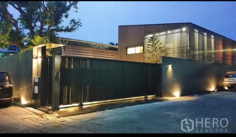 Home Office for rent or sale in Khlong Toei, Bangkok - Modern gated home office exterior with tall black fence, warm uplights and a tree-lined street at dusk in Phra Khanong.