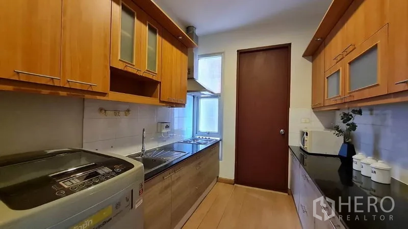 Condominium for rent in Khlong Toei, Bangkok - Galley kitchen with wooden cabinetry, built-in cooktop, range hood, window, and washing machine.