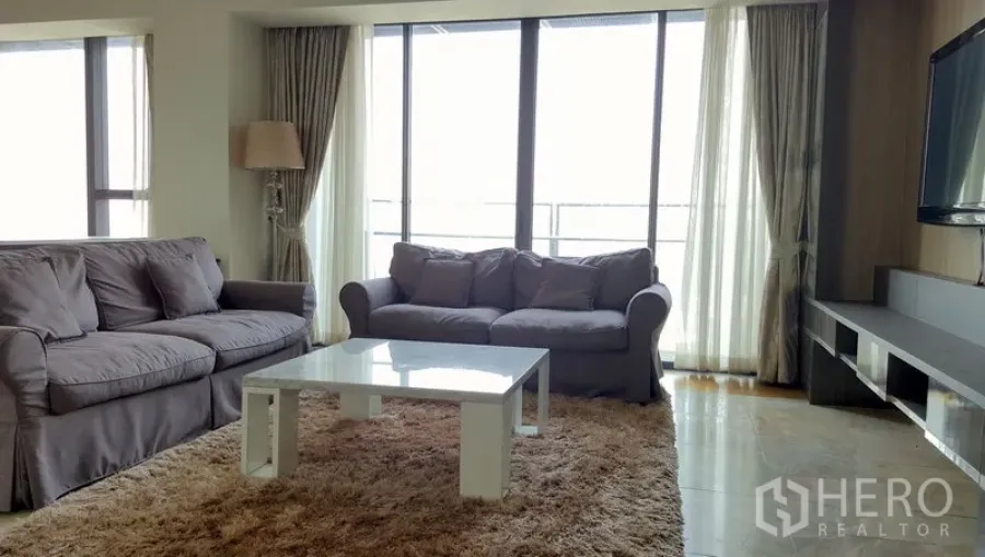 Condominium for rent in Sathon, Bangkok - Bright living room with two gray sofas, shaggy rug, white coffee table, and floor-to-ceiling windows opening to a balcony.