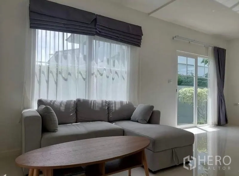 Detached House for rent in Bang Bo, Samut Prakan - Bright living room with L-shaped gray sofa, sheer curtains and a sliding glass door to the garden.